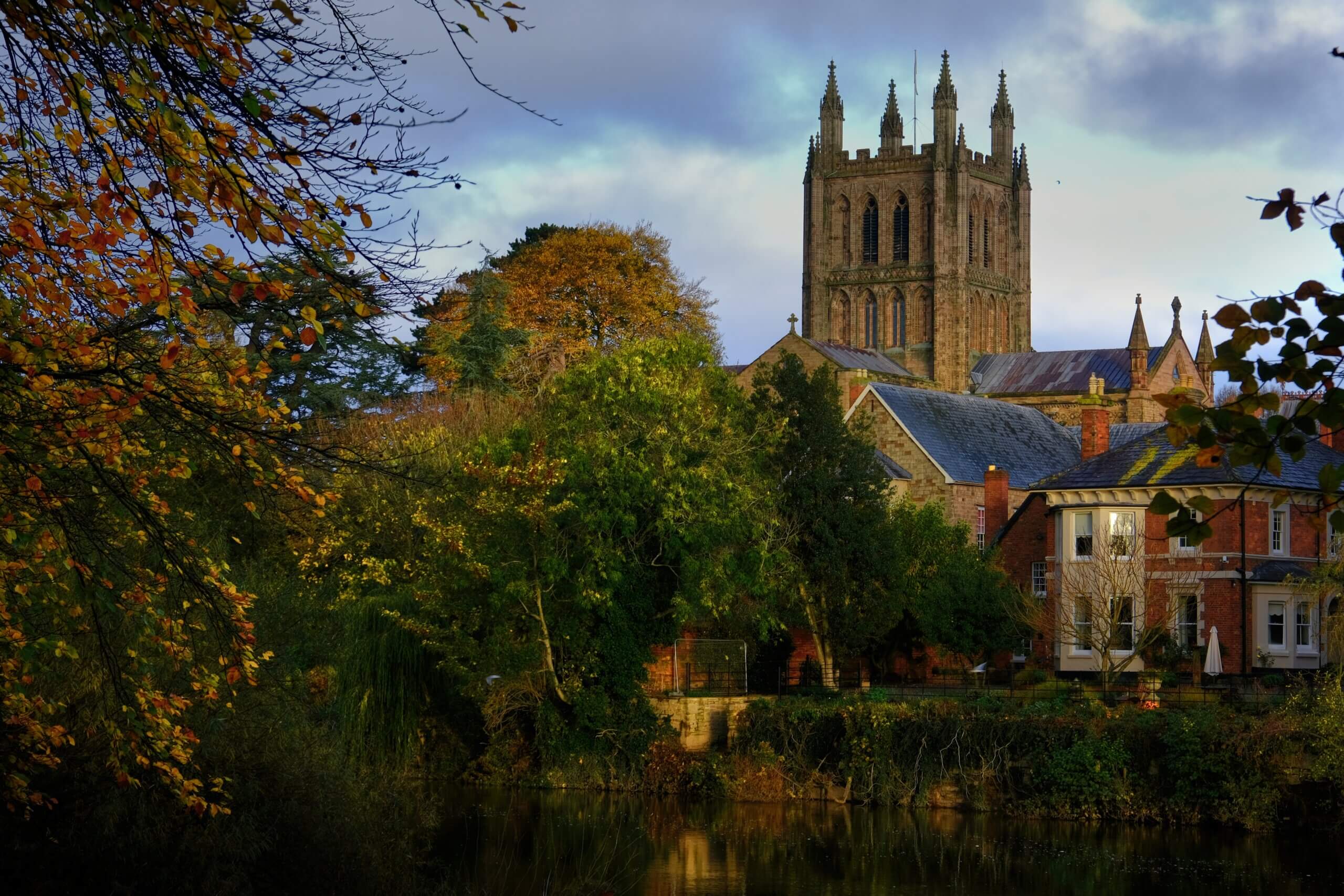 An image of Hereford Cathedral surrounded by trees and a river.